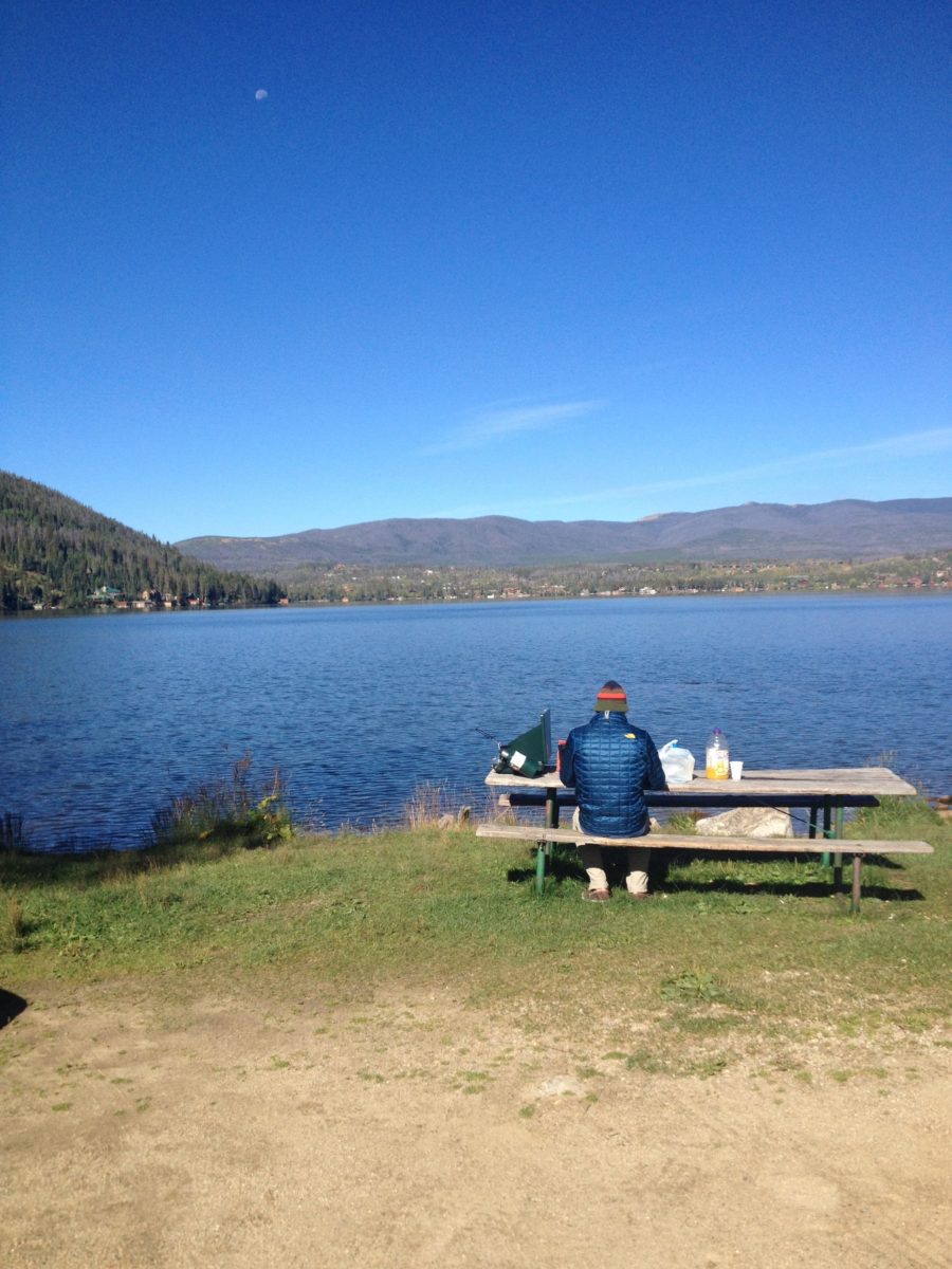 Man sitting at picnic bench near a lake on a blue sunny day.