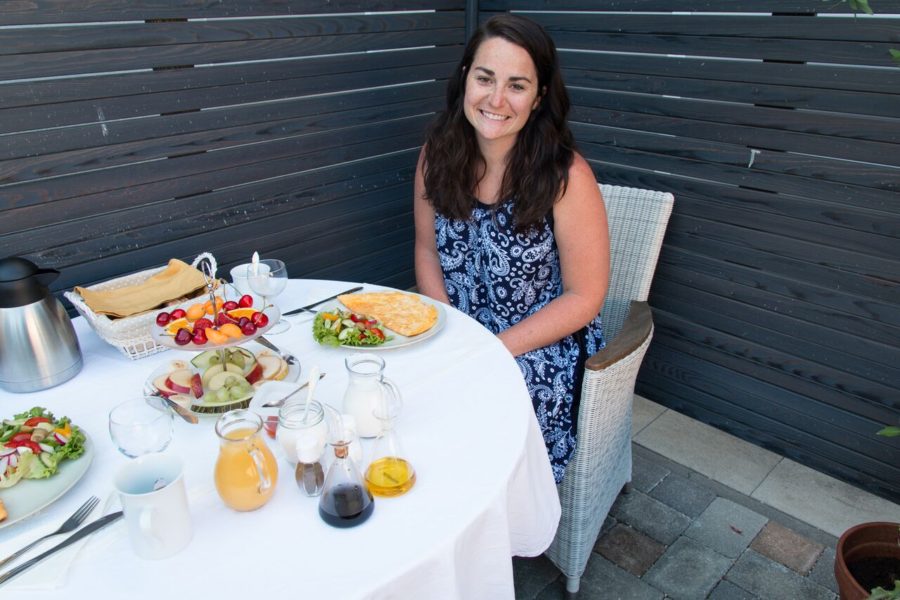 breakfast spread on table with white tablecloth and woman sitting in chair smiling at camera