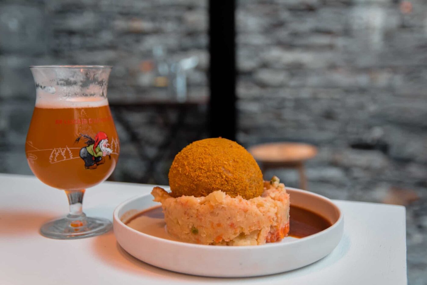 breaded ball on mashed potatoes next to a belgian beer on a table at a restaurant in belgium.