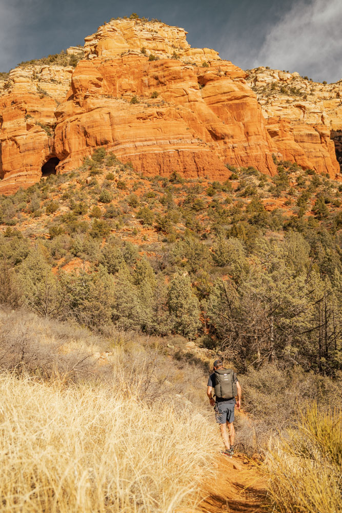 A man walking on a small social trail in Sedona with a backpack full of camera gear, as he hikes towards the red rocks.