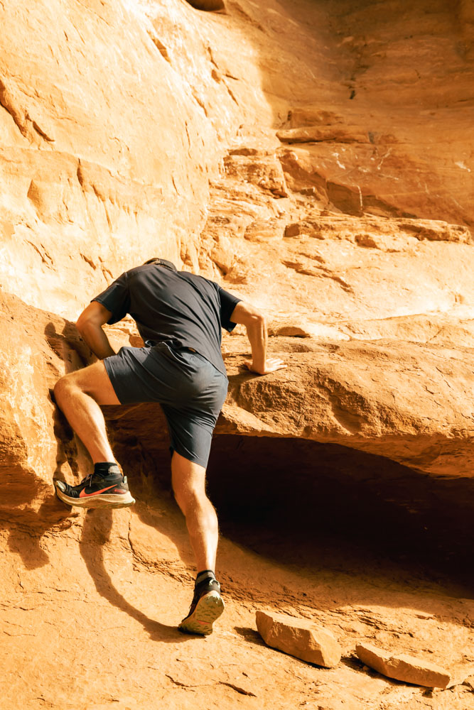 Man in shorts and t-shirt wearing extremely grippy shoes climbing up into a cave in Sedona.