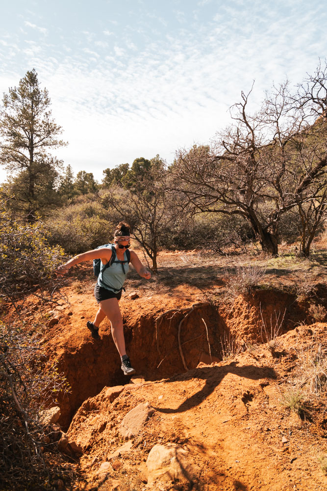 Woman hiking in blue tank top and black shorts jumping over a wash in Sedona on her way to Keyhole Cave.