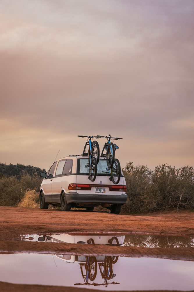 A Toyota Previa campervan with bike rack and van reflecting off rain puddle in foreground.