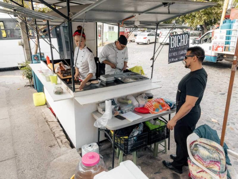 Man in black clothes standing outside street taco stand in Puerto Vallarta with workers inside in white chef's outfits.