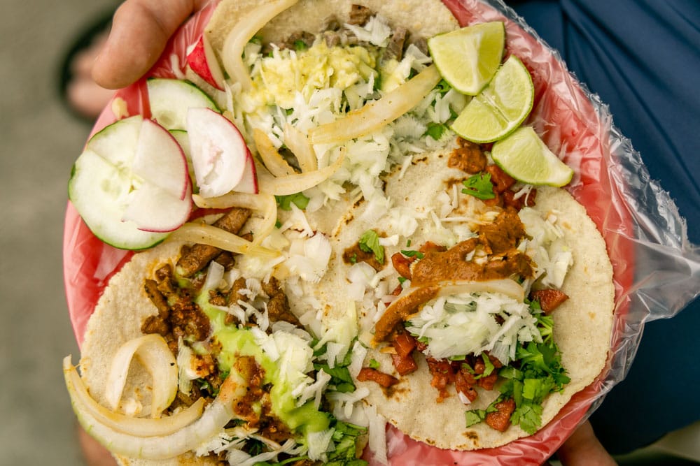 A red plate with plastic bag over it, and three street tacos on top with limes, radish, and cucumber.