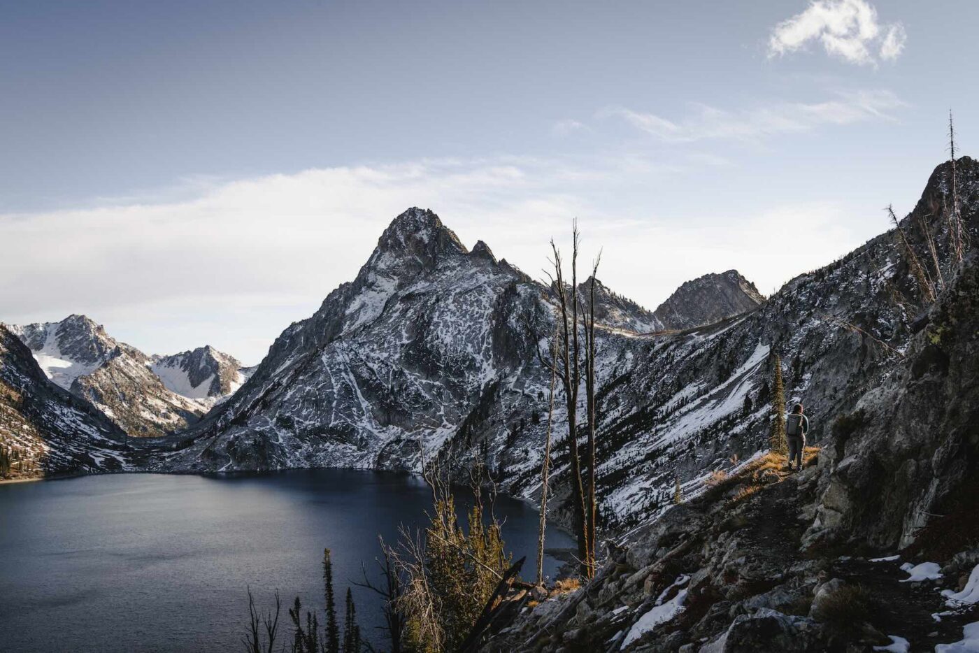 A lone hiker, Heather, with a backpack walks along a rugged mountain trail, surrounded by snow-dusted peaks and a deep alpine lake. The towering, jagged mountains dominate the landscape under a clear blue sky, with patches of sunlight casting a golden glow on the slopes. The scene captures the essence of remote wilderness and adventure.