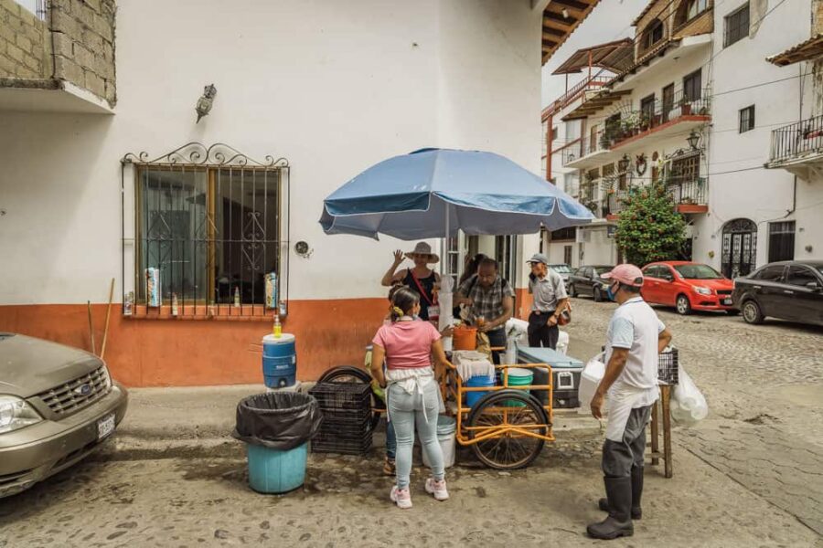 Small push cart taco stand with blue umbrella and woman waving at camera from it.
