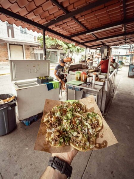 An arm holding out a plate of seven small tacos in front of a taco stand in Puerto Vallarta, Mexico.