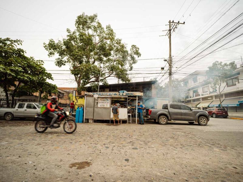 A motorcycle driving by a taco stand with smoke from the grill coming out on the street in Puerto Vallarta.