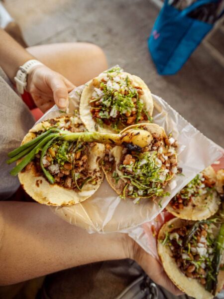 A woman holding two plates with three street tacos on each plate and a grilled spring onion on top.