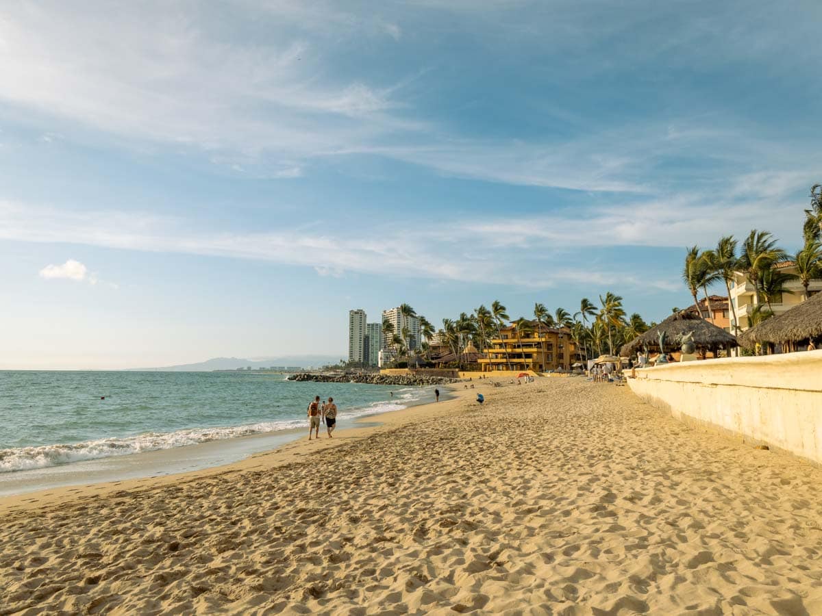 A vacant beach in Puerto Vallarta with couple walking along the shore and tables and chairs in the distance.
