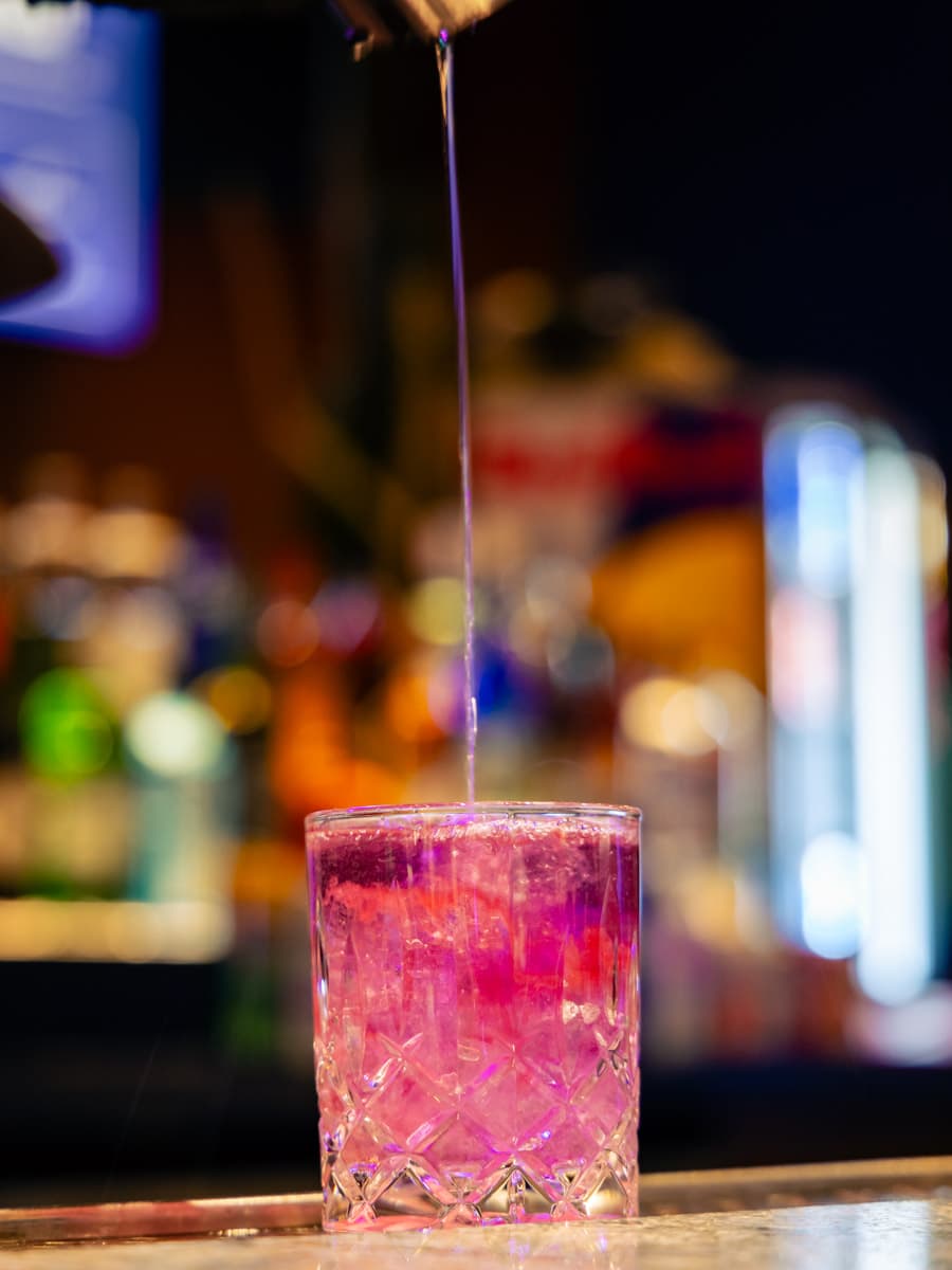 A pink drink at a bar with drink being poured from high above.