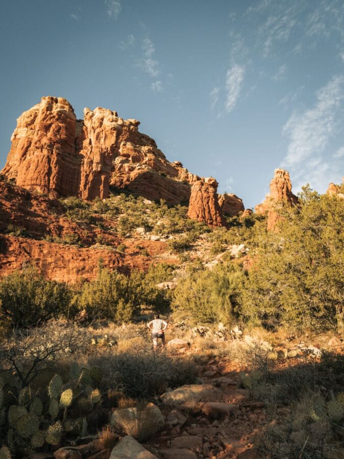Trail finding in Sedona with bushes and cacti on both sides of trail and red rock formations in site.