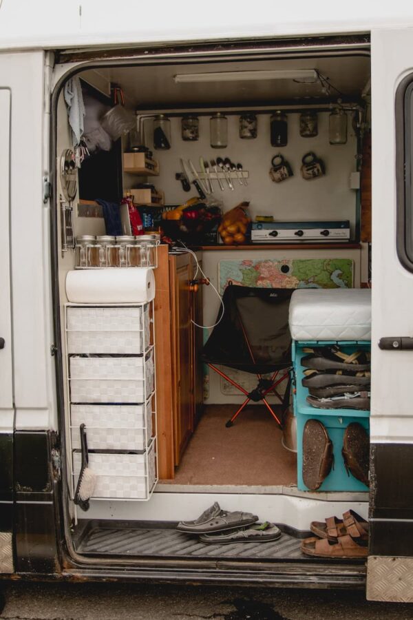 Looking inside a campervan conversion with hanging jars holding dry foods, a white drawer basket shelf and stove on top of counter below the hanging jars.