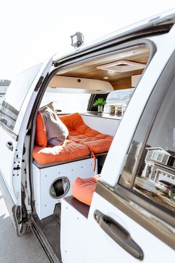 Looking inside a minivan campervan conversion with orange cushings as the bed and white paint for the wood with a cat hole by the door.
