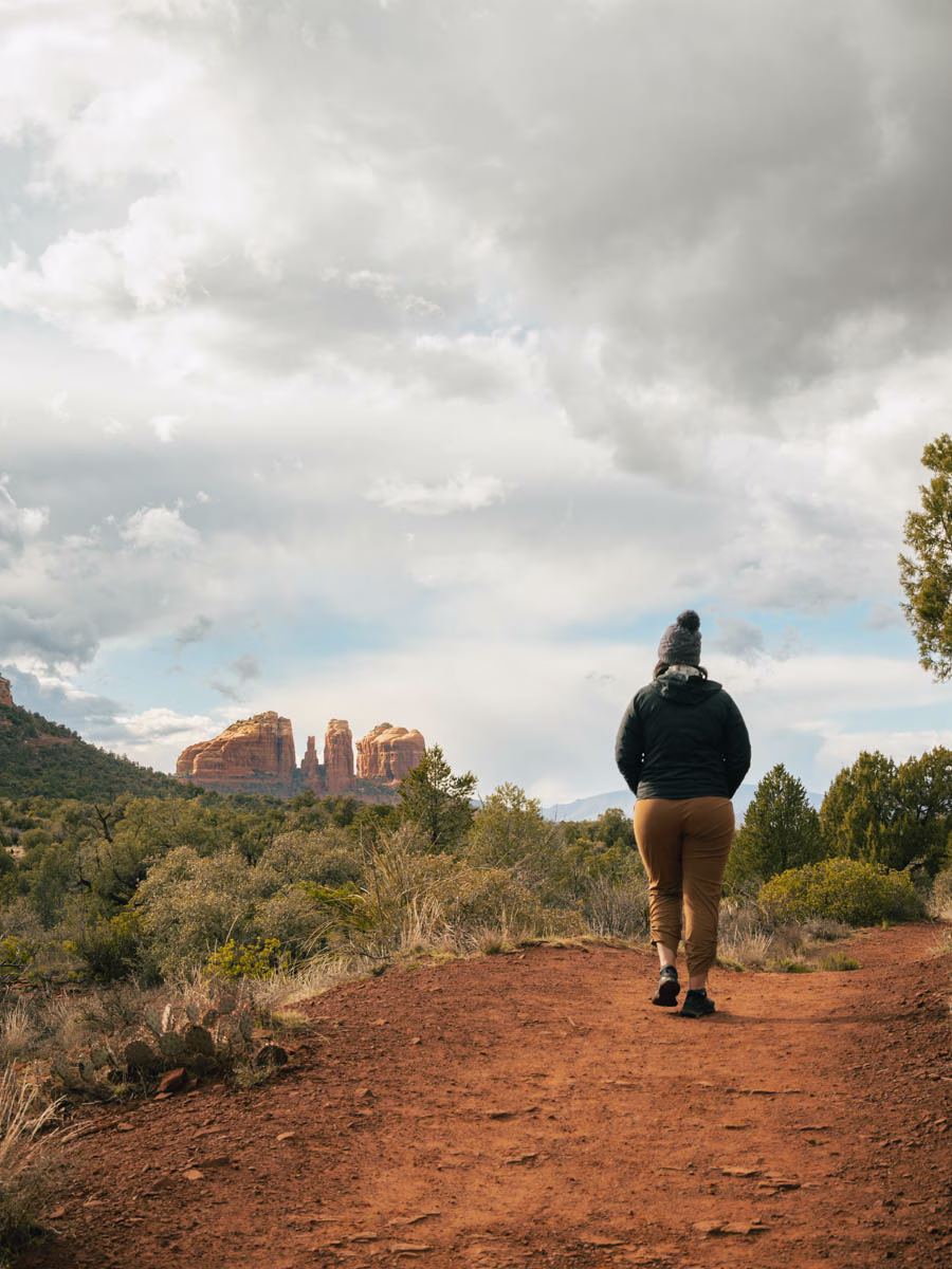 A woman on an easy hiking trail in Sedona on red dirt and rock looking out towards Cathedral Rock.