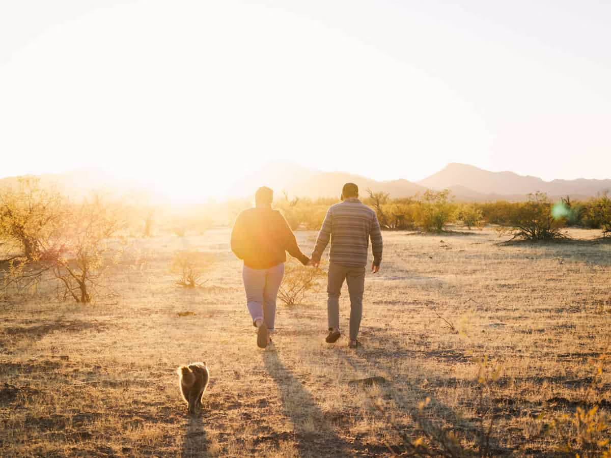 Married couple, Heather and Jason, hold hands walking into the sunet with cat, Georgie, following closely behind.