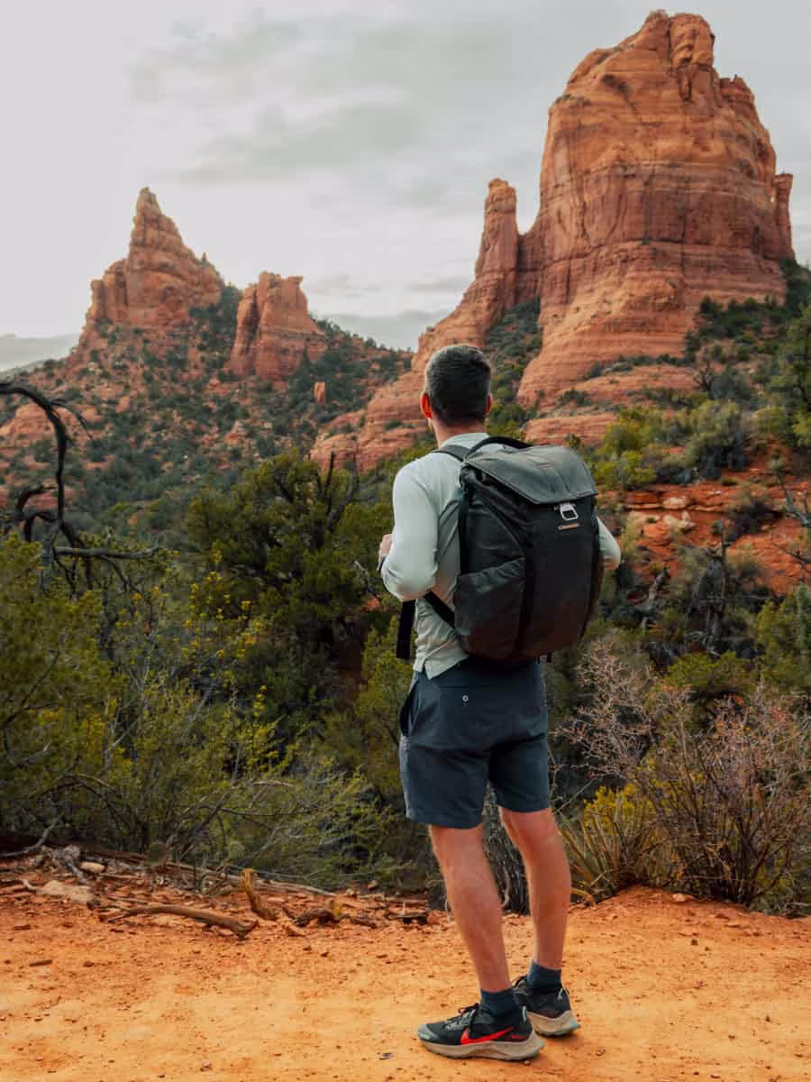 A male hiker wearing a black backpack stands on a red dirt trail, looking at towering red rock formations surrounded by desert vegetation under an overcast sky.