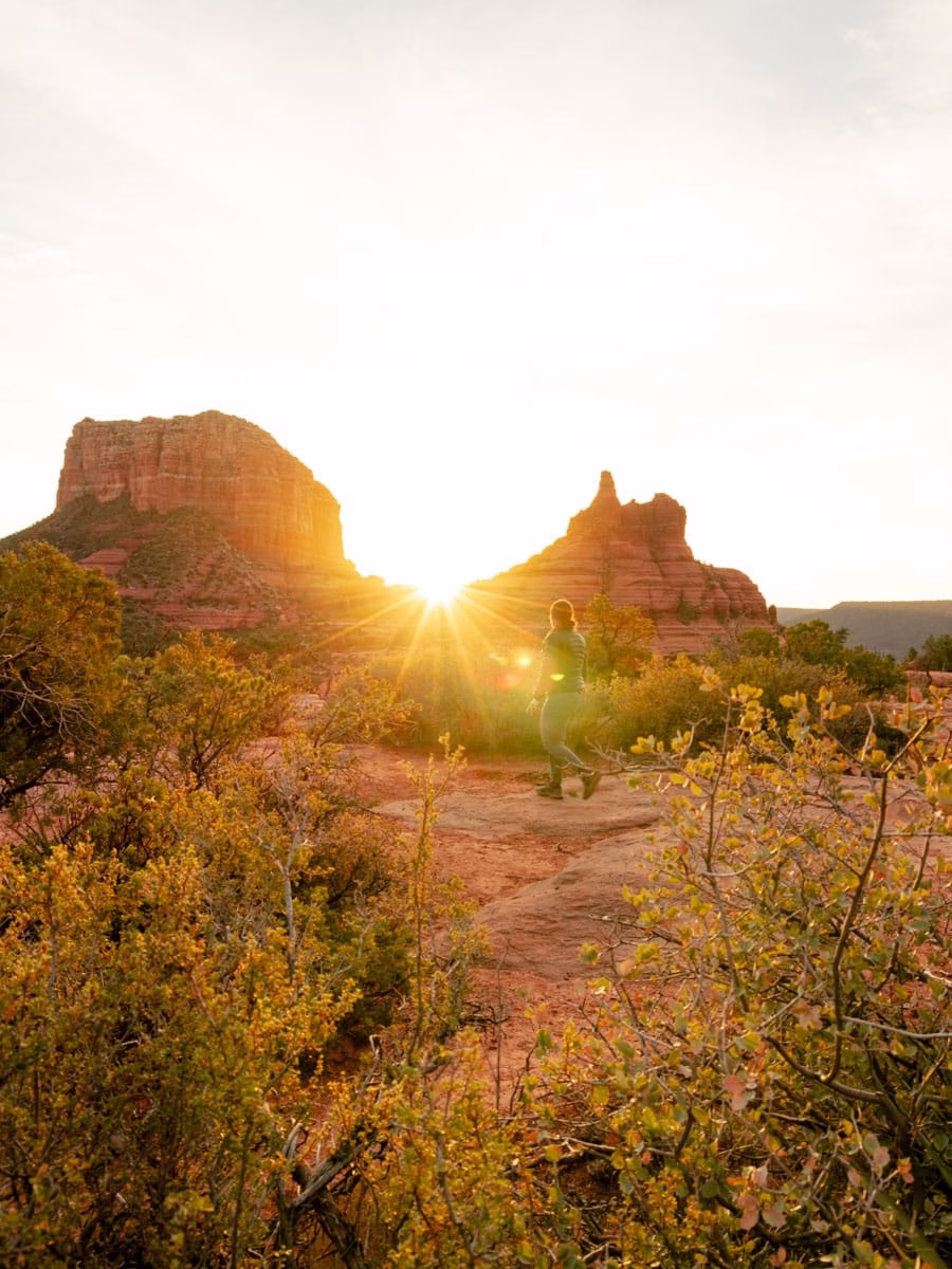 Sunrise peeking between Courthouse Vista and Bell Rock with woman walking across the frame.
