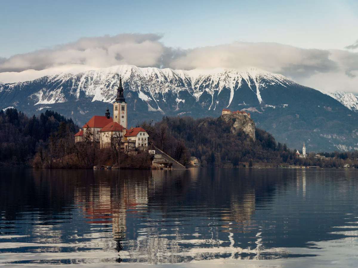 The Lake Bled cathedral on the left side of the photo, with the castle a in the distance to the right, and snow covered mountains towering them both in the background.